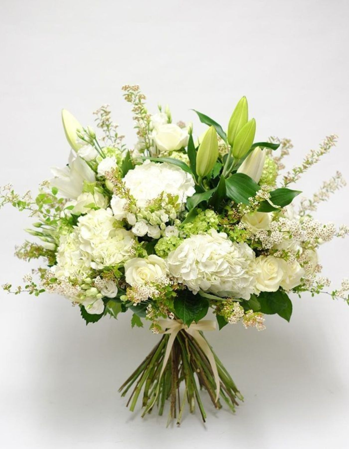 A white and green floral bouquet featuring hydrangeas, lilies, roses, and filler flowers, tied with a beige ribbon, set against a plain light gray background.